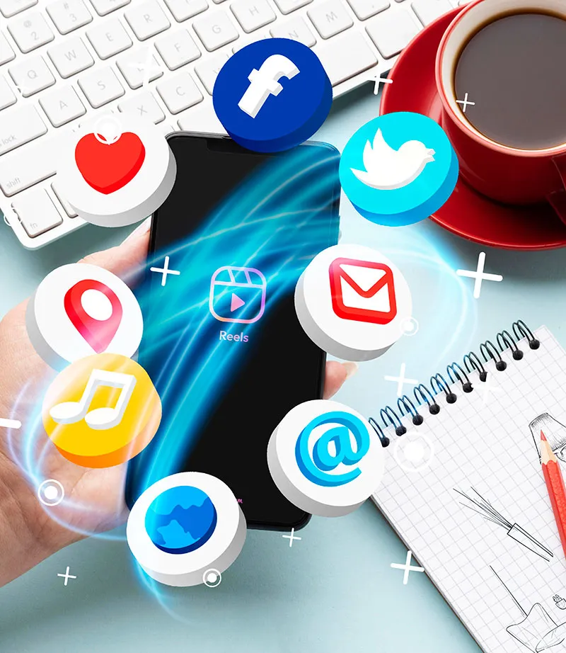 A hand holds a smartphone displaying app icons for social media, email, and music, with a keyboard, coffee cup, notepad, and pen visible on the desk.